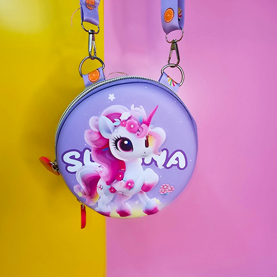 Two colorful, round kids’ sling pouches with attached lanyards are placed on a sandy beige background with a tropical leaf shadow. The left pouch is purple and features an adorable cartoon unicorn with a pink mane, large sparkling eyes, and the name "SHYAMA" written in white. It has a lilac lanyard decorated with candy prints. The right pouch is bright blue with a fierce green cartoon T-Rex dinosaur illustration labeled “DINOSAURS” in bold yellow text. Its lanyard is blue with black dinosaur silhouettes. Bo
